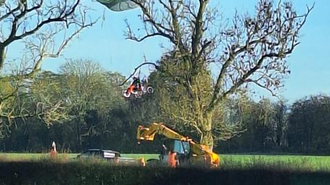 A glider stuck in a tree, with a yellow crane below. There is a man in the glider.