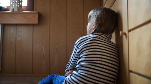 A young girl in a striped jumper and jeans with her hair in bunches sits with her back to the camera looking up at a window
