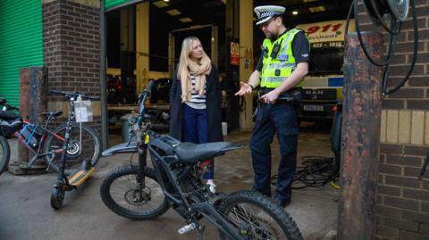 BBC reporter Hope Webb, wearing a blue jacket and cream-coloured scarf, stands next to police inspector Hugh Niccolls, who is wearing a green fluroescent police vest. They are both standing next to a large, grey e-bike which resembles a dirt bike parked in front of a warehouse entrance.