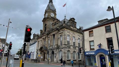 Kendal's historic town hall, with a clock tower, situated on the streets in the centre of the town and surrounded by busy roads and shops.