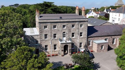 A drone shot of Alderney Island Hall. It is a grey-stone Georgian building with long windows and a large porch area. The building is surrounded by trees.