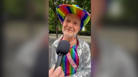 Keith, 64, wearing a rainbow cowboy hat, rainbow vest and silver sequined jacket, speaks into a microphone.