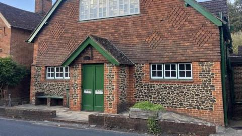 A small brick and tiled village hall with a green door. It is beside a road in a village