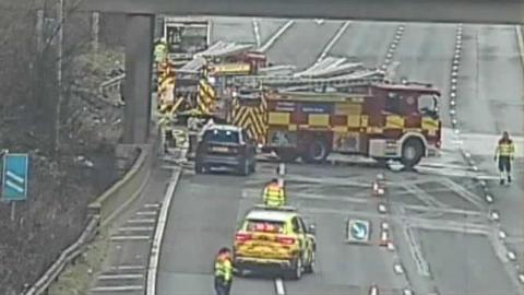 The M25, with several lanes of traffic, with police vehicles, fire engines, and people standing on the road, in high-vis jackets. There are cones on the road.