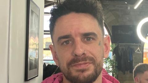 A man with short brown hair wearing a pink hoodie stands in a barber's shop. He has a tattoo on his left cheek and two nose piercings.