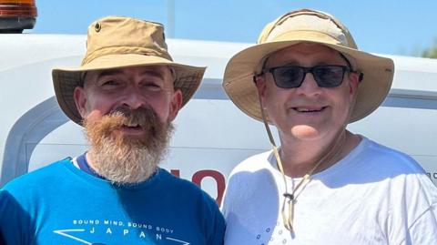 Two men wearing hats. One has a blue t-shirt on and one has a white t-shirt on. A blue sky is behind them. 