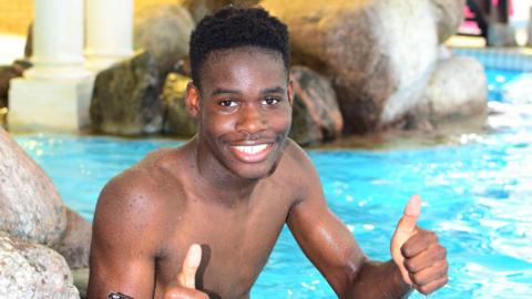 A young man smiling with his thumbs up. He is topless and there is a swimming pool behind him, with some rocks along the side of it.