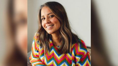 A woman with shoulder length, wavy brown hair smiles for the camera. She is wearing a multicoloured, zig-zag patterned knitted jumper.