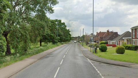 A long straight residential road with a white line down the middle. There are green tress lining the left hand side of the road. On the right are a number of houses with green lawns in front of them.