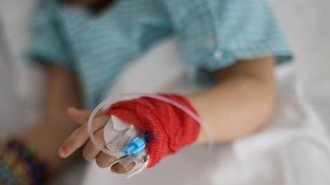 Close-up of a patient's hand wrapped in a red bandage with an intravenous (IV) cannula inserted and tubing attached, resting on a hospital bed. The person is wearing a light blue hospital gown, with the background softly out of focus.