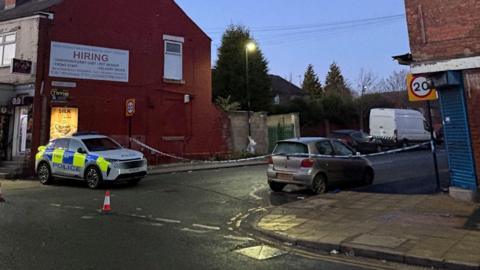 A street at dusk showing a police car parked on the left side near a red brick building with a large white sign. The area is cordoned off with police tape, and traffic cones are placed on the road. A silver car is parked on the right side of the street, and a white van is visible further down the road. There are 20 mph speed limit signs on both sides of the street.