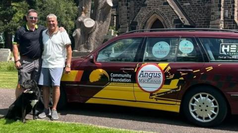 Two men and a dog stand to the left in front of the bonnet of a large, burgundy estate car with logos all over it. The entrance to a church is visible right behind the car.