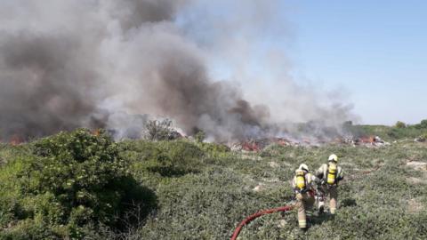 A fire burning at Arnolds Field in east London. You can see two firefighters approaching the blaze with a hose. 