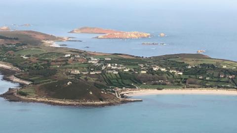 An aerial photo of St Mary's on a sunny day. The island has fields and houses and a long white sand beach can be seen. There are further small islands beyond.