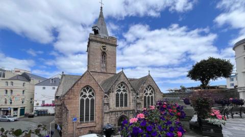 A granite church with a slightly reddish hue, set among a shopping street. Purple and pink flowers in the foreground and a blue, cloudy sky.