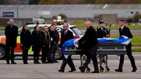 Alex Salmond's coffin, coverd in a Saltire, is being carried across the tarmac by funeral staff on a trolley. Friends and family are standing in a line in the background