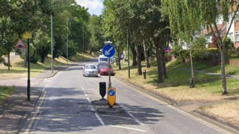 StreetView image of a road island and signs on Watling Street near the junction with Halcot Avenue