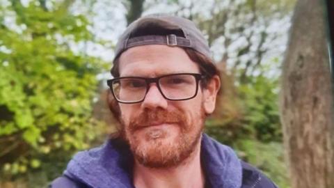 Shane Lynch with a hat on and glasses, Shane has a beard and is standing by some trees.