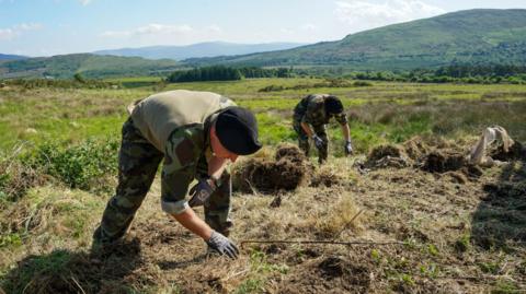 Two people wearing army clothing are digging in the ground with there hands. They are wearing gloves. In the background we can see a collection of hills and rugged countryside land. 