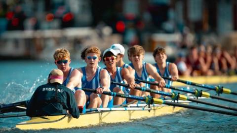 Crew of eight in blue striped race suits mid-race onboard a yellow rowing boat