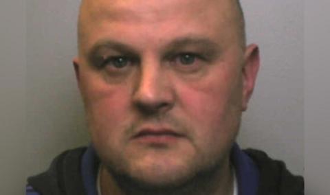 A police mugshot of a man with a shaved head, stubble and blue eyes. A blue and black hoodie and neck of white t-shirt can just be seen.