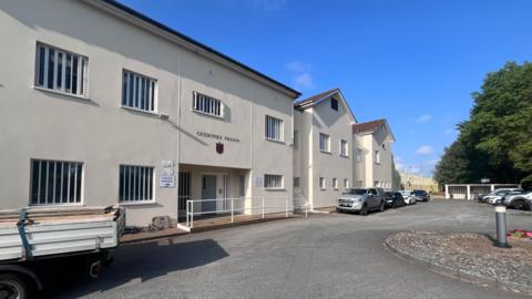 This is an exterior shot of Guernsey Prison. It is a cream building with a ground and first floor. There is a driveway with cars parked in front of it. There are tall trees behind them, and the sky is bright blue.