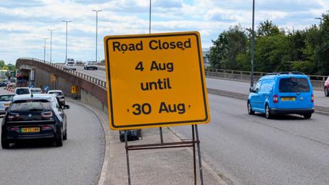 A view of the flyover with cars driving over it. Several drive to the side of it. A yellow sign in the middle says "Road closed - 4 Aug until 30 Aug".