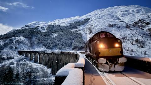 A train enging, with snow plough attached, crossing a Highlands viaduct in a snowy landscape. The sky is blue and the train is coming towards us with lights on.
