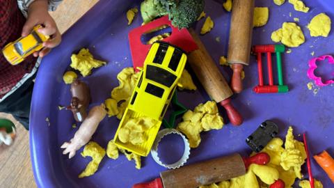 Clai gyda anifeiliaid a siapaiau wedi eu gosod ar fwrdd porffor gyda dwylo plentyn yn ymestyn am y tegannau. 