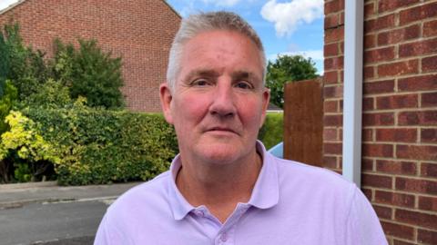 Mark Hemphill stood on a residential street. He is wearing a purple polo tshirt and is staring into the camera.