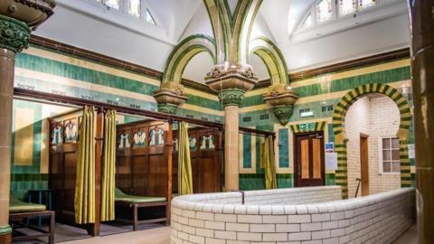 Carlisle's Turkish Baths, pictured before the closure. The walls are tiled in green and white. There are three small, curtained cubicles to the left with wooden benches in each one.