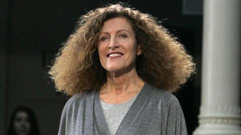This is a photo of a woman with brown curly hair, wearing a grey top and smiling at the camera. There is woman standing in the distance behind her.