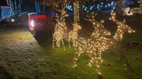 Six illuminated reindeer sculptures are styled to appear to be towing a red sleigh. They are in a garden with hedges and trees also illuminated.