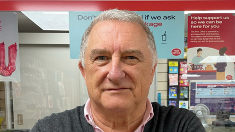 An elderly man standing in front of a Post Office kiosk staring in the camera