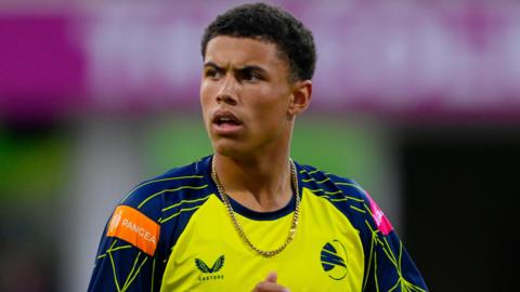 Head and shoulder shot of Hampshire bowler Manny Lumsden looking to his right, wearing a Hawks T20 yellow and blue kit and a gold chain