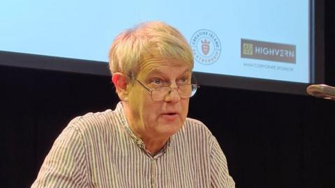 A man, in a white and purple striped top. He is looking forwards and his mouth is slightly open. He is wearing glasses. Behind him is a projector screen and black curtain. 