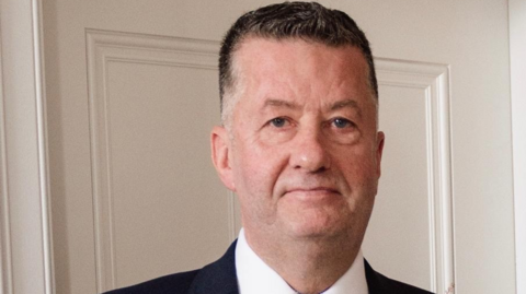 Jim Wylie, a man with short, dark hair which is greying at the sides. He is wearing a dark three-piece suit, a white shirt, a silver tie and a wedding buttonhole on his lapel