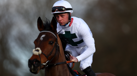 Levi Williams sitting on a brown horse, wearing an all-white outfit, with a white helmet and black glasses. He is holding on to the reins of the horse.