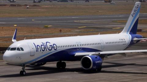 A white aeroplane with the word 'Indigo' on its side in blue writing stands on a runway. 