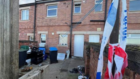 The exterior of a row of terraced houses. The patio out front is littered with appliances, including cookers, and police tape is tied to a pole in the foreground. No fire damage can be seen on the outside of the house but the bottom window is boarded up.