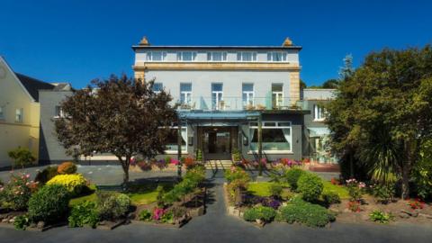 The hotel is grey with a balcony on the second floor. It has two grass patches with flowers and trees in front.