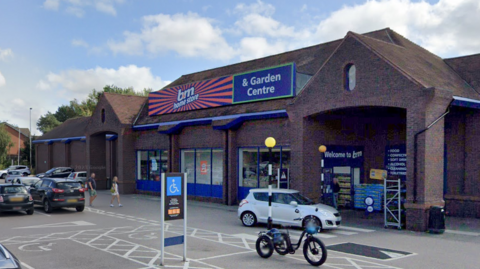 The car park and entrance to B&M supermarket on the outskirts of Ipswich. The orange and blue branding on the building is visible. A few cars and people are also seen