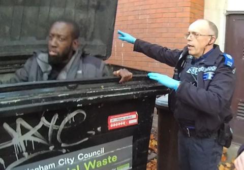 Ashley Lawrence, who has short dark hair and a beard sitting up in a bin, with a police officer who has short hair stood next to the bin wearing uniform and blue latex gloves.