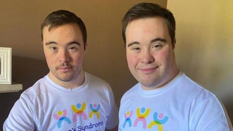 Two young men are smiling as they stand side by side in a house, both wearing white T-shirts with the pink, yellow and blue Down's Syndrome Manchester logo on.