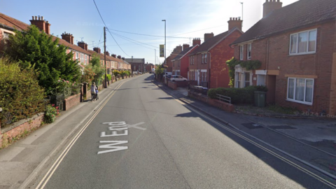 A road with houses on either side and cars passing in the distance.