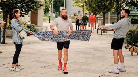 Joe smiles as he runs through the finish tape. He's wearing a white running top and people in the background are taking photos and clapping him. His hair is tied into a bun at the back of his head and his hair is shaved on the sides. He has a long beard as well.