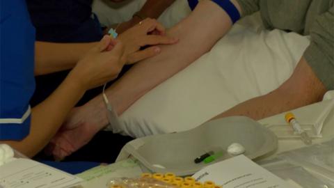 A patient is about to receive a blood test. A nurse is holding a needing in one hand and has placed her finger of the vein on a man's arms. The man is sitting down and is resting his arms on a pillow in a hospital setting. 