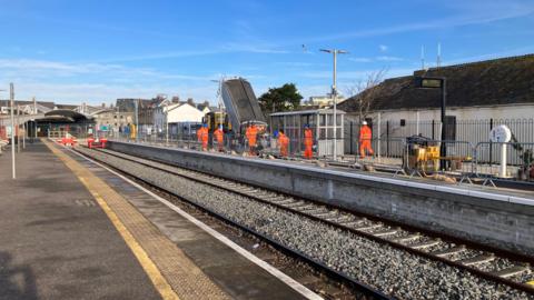 A photo from the side of a station platform where workers are working on the other side of the platform. It's the end of the railway line with two tracks. 