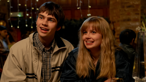 The main characters Owen and emily sit side by side at a wooden bar table in a warmly lit, busy pub or bar. Both lean slightly forward with their arms resting on the table. Owen wears a light-coloured jacket over a checked shirt, while Emily wears a dark jacket, with blonde hair falling around her shoulders.