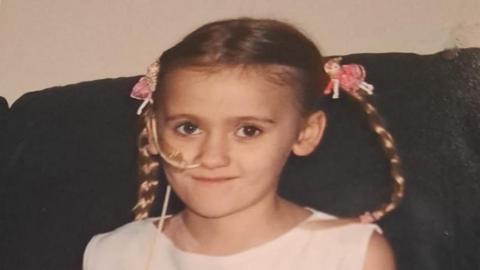 A young girl with her blonde hair in braided pig tails. She has a white tube up her nose.